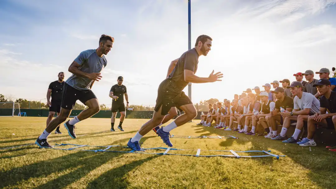 Players running training camp drills as fans watch from a sunny berm