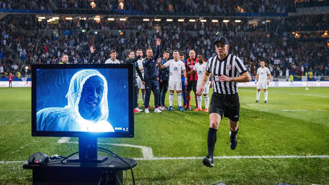 A referee reviews a play at the pitch-side VAR monitor as players crowd behind in suspense