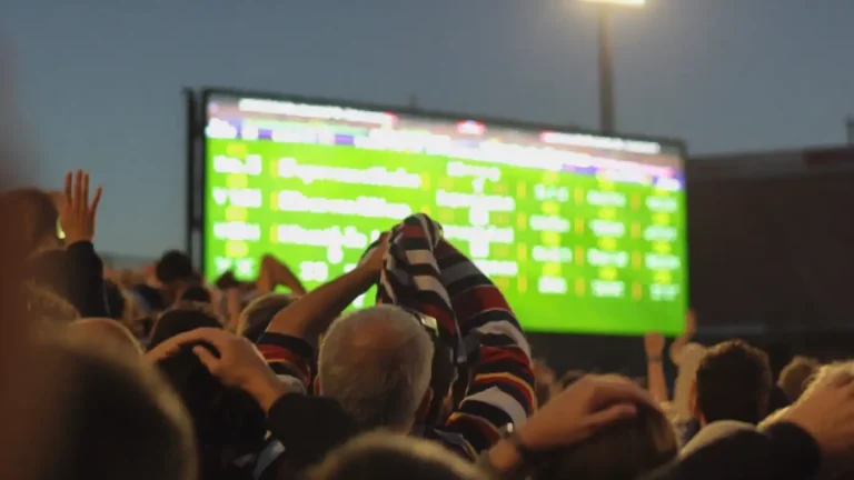 Anxious fans staring up at a giant stadium scoreboard at dusk with hands on heads